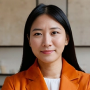 Portrait of a smiling Asian woman wearing an orange blazer and white top against a beige background.