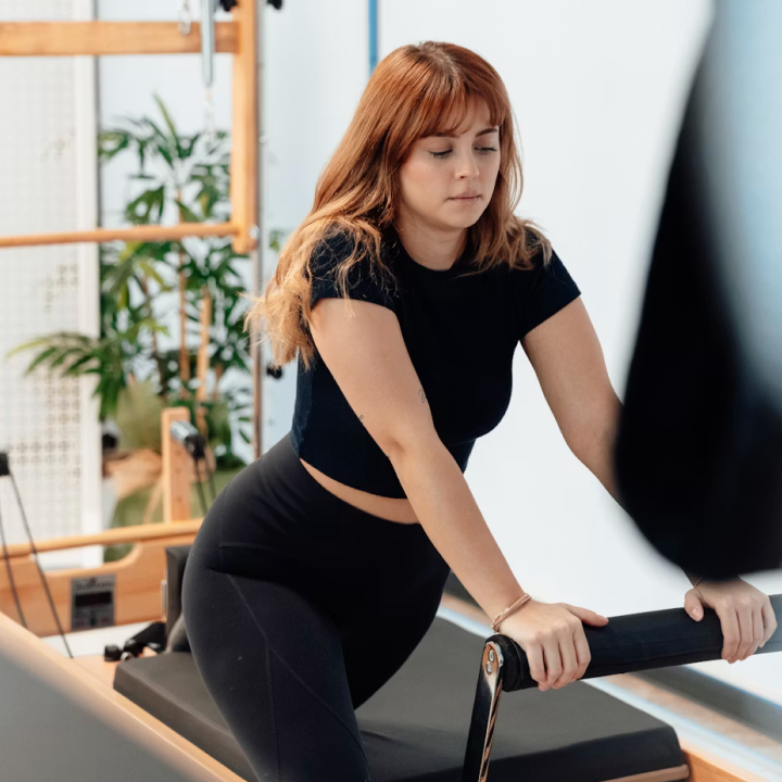 Young woman in black workout clothes using Pilates reformer machine indoors with plants in background.