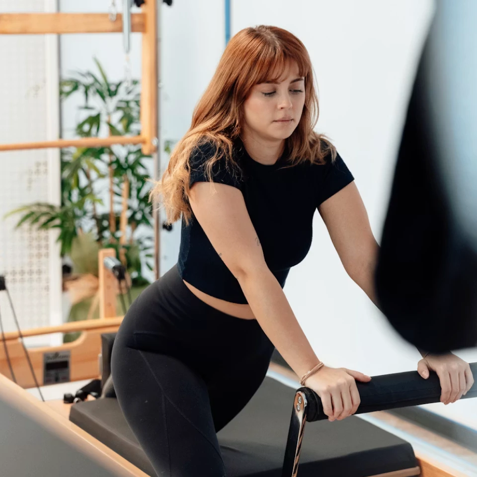 Woman with red hair in black workout clothes using Pilates reformer machine indoors.