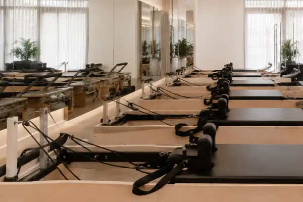 Row of Pilates reformer machines in a bright studio with mirrors and plants near large windows.