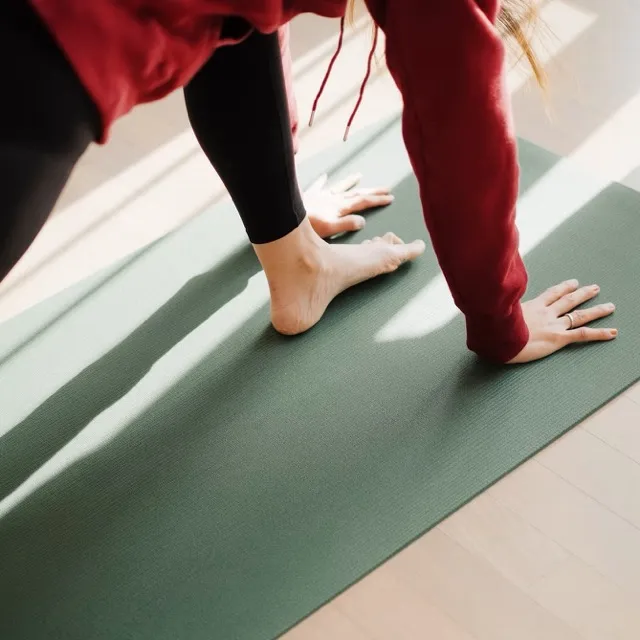 Person wearing red hoodie and black leggings doing yoga stretch on green mat in sunlight.