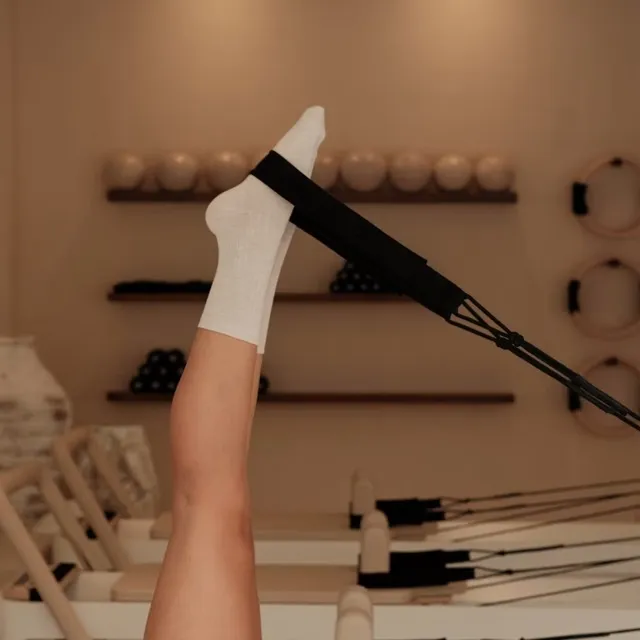 Leg extended upward with foot in white sock strapped in Pilates reformer exercise machine.