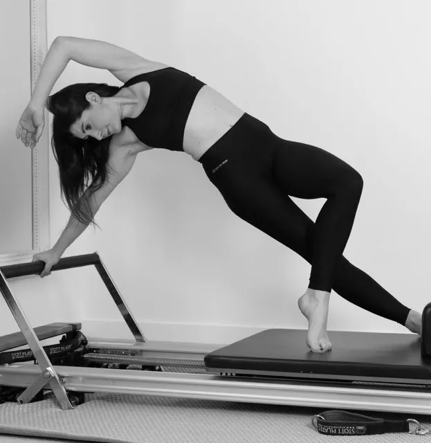 Woman in black workout clothes doing a side plank on a Pilates reformer machine.