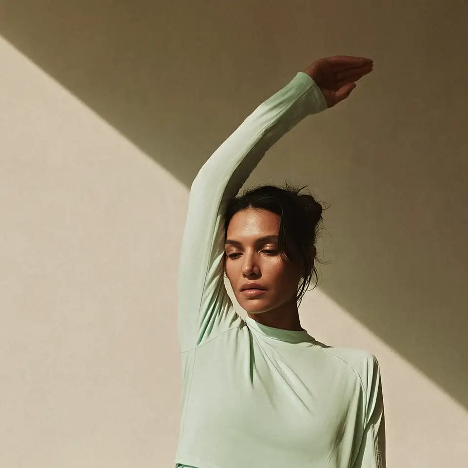 Woman in light green top with one arm raised against a beige wall with diagonal shadows.