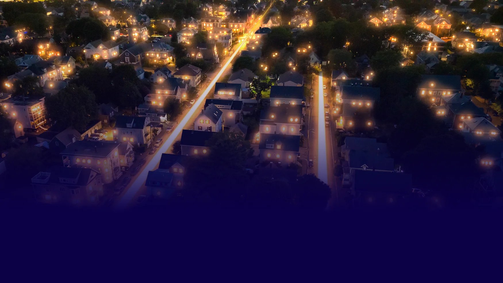Night aerial view of a residential neighborhood with streetlights and warm lights in house windows.