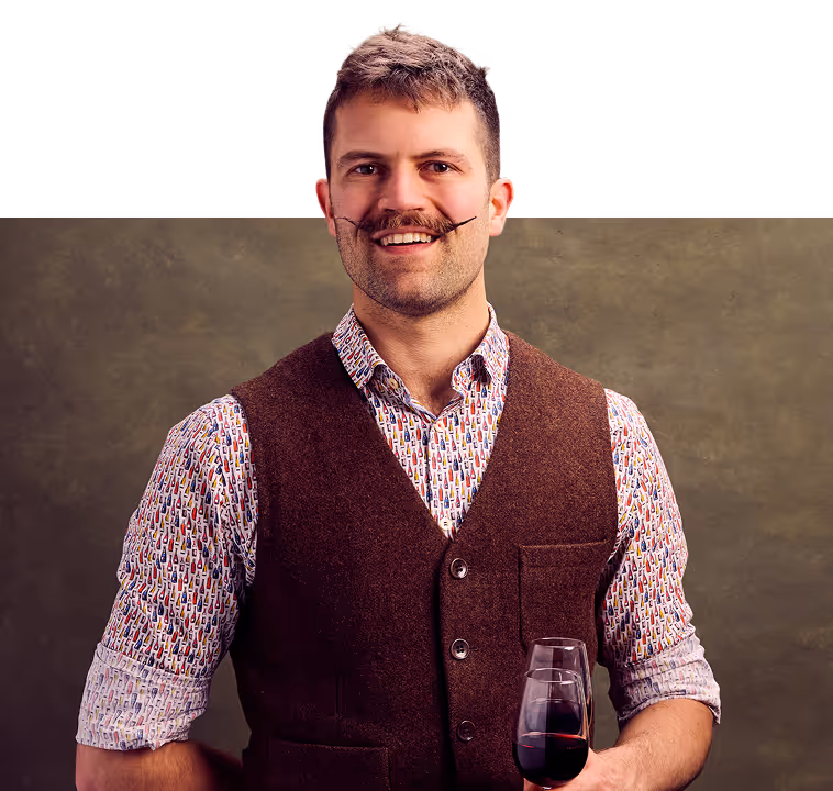 Smiling man with a styled mustache wearing a patterned shirt and brown vest, holding a glass of red wine.