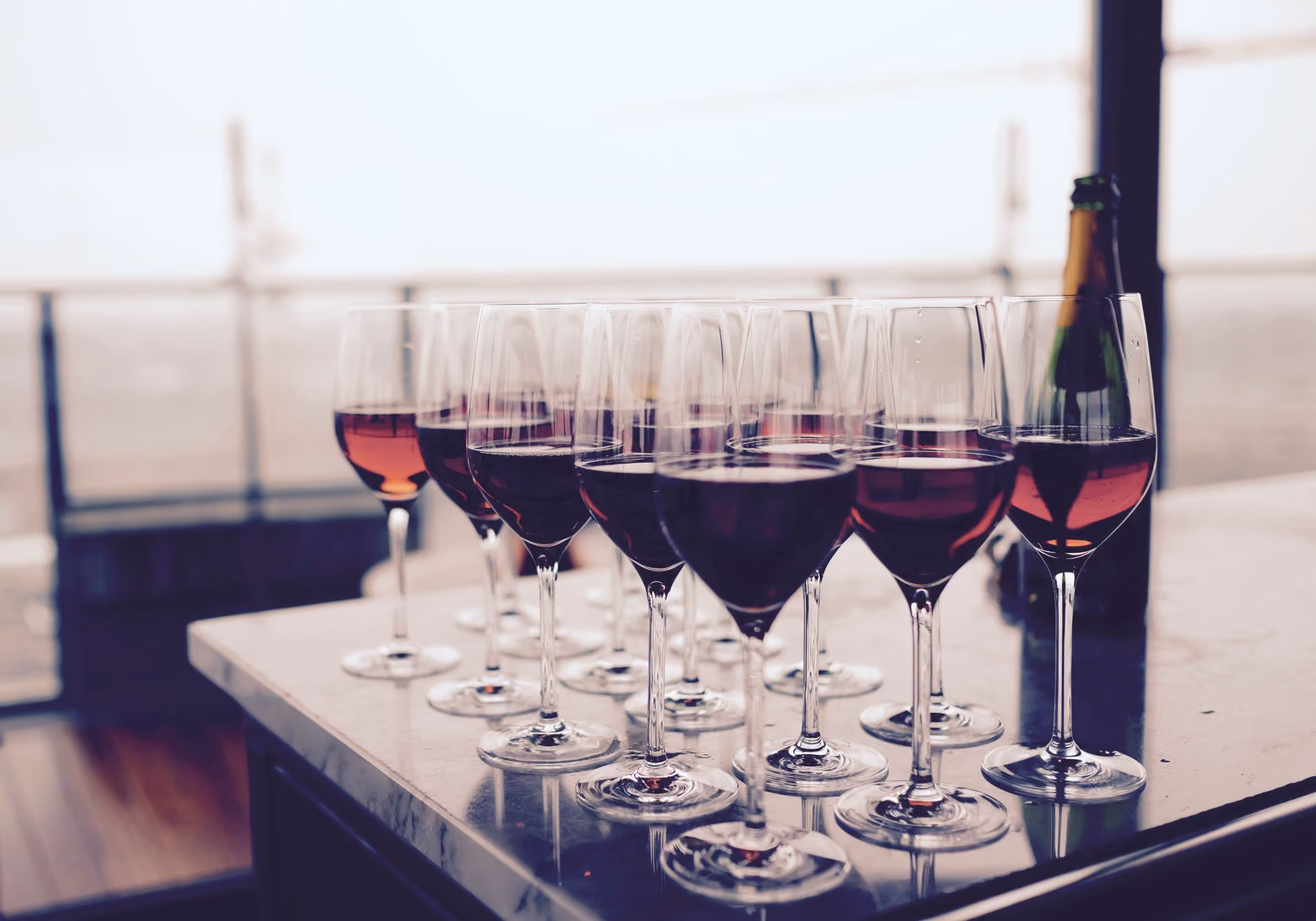 Multiple wine glasses filled with red wine arranged on a marble table with a bottle in the background.