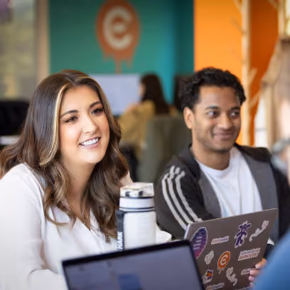 Two young adults sitting at a table with laptops, smiling and engaged, in a colorful indoor setting with teal and orange walls.