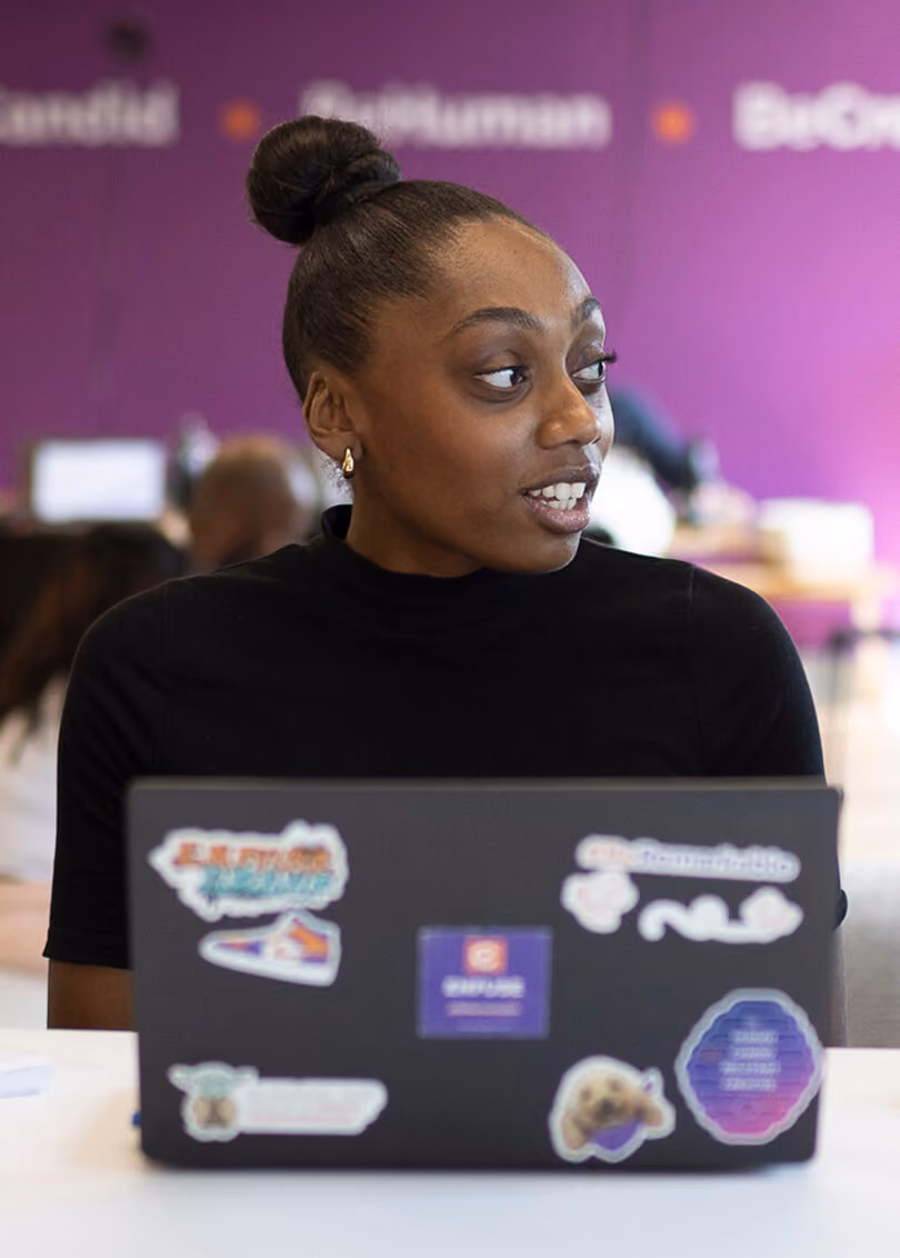 A woman with a high bun hairstyle and gold earrings is sitting at a table with a laptop covered in colorful stickers, looking to her right. The background is blurred with a purple wall and indistinct people and objects.
