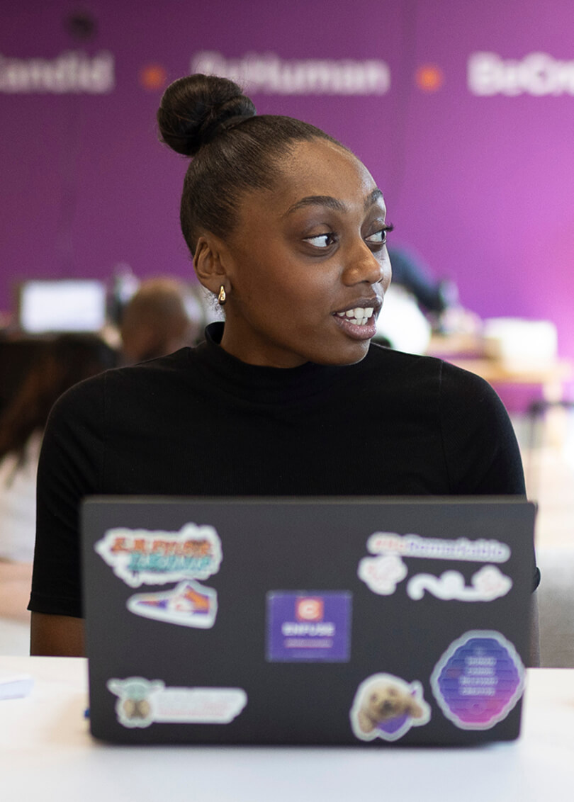 A woman with a high bun hairstyle and gold earrings is sitting at a table with a laptop covered in colorful stickers, looking to her right. The background is blurred with a purple wall and indistinct people and objects.