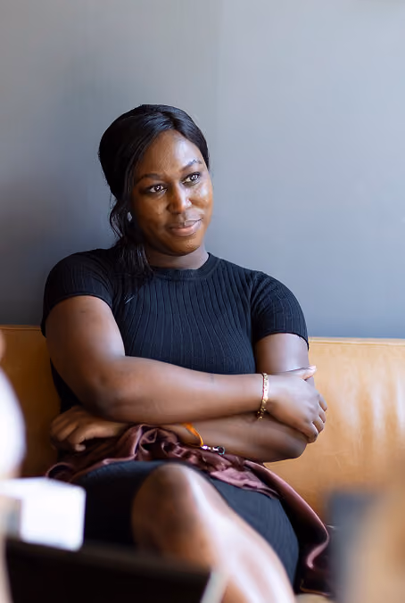 A woman with dark hair tied back, wearing a black ribbed short-sleeve top, sitting on a tan leather couch with her arms crossed, looking thoughtfully to the side against a plain gray wall.