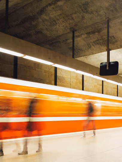 Blurred figures walking on a subway platform with an orange and white train passing by, under a concrete ceiling with lights.
