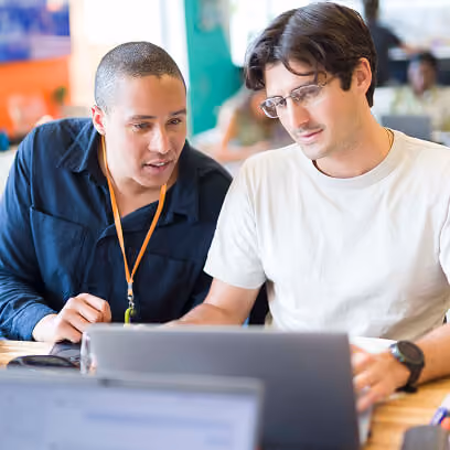 Two men sitting at a table working together on a laptop, one wearing glasses and a white shirt, the other wearing a dark jacket with an orange lanyard.