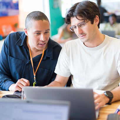 Two men sitting at a table working together on a laptop, one wearing glasses and a white shirt, the other wearing a dark jacket with an orange lanyard.