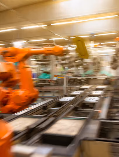 Orange industrial robotic arm operating on an assembly line in a factory setting with blurred background of machinery and workers.