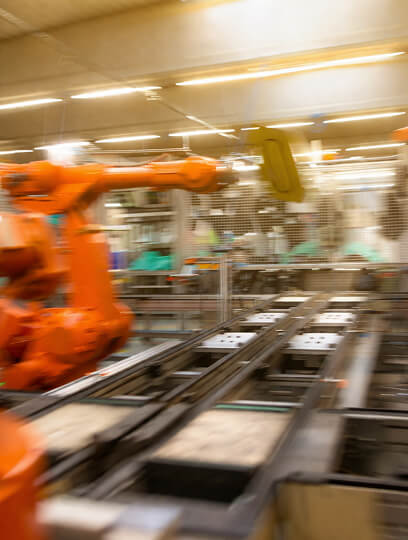Orange industrial robotic arm operating on an assembly line in a factory setting with blurred background of machinery and workers.