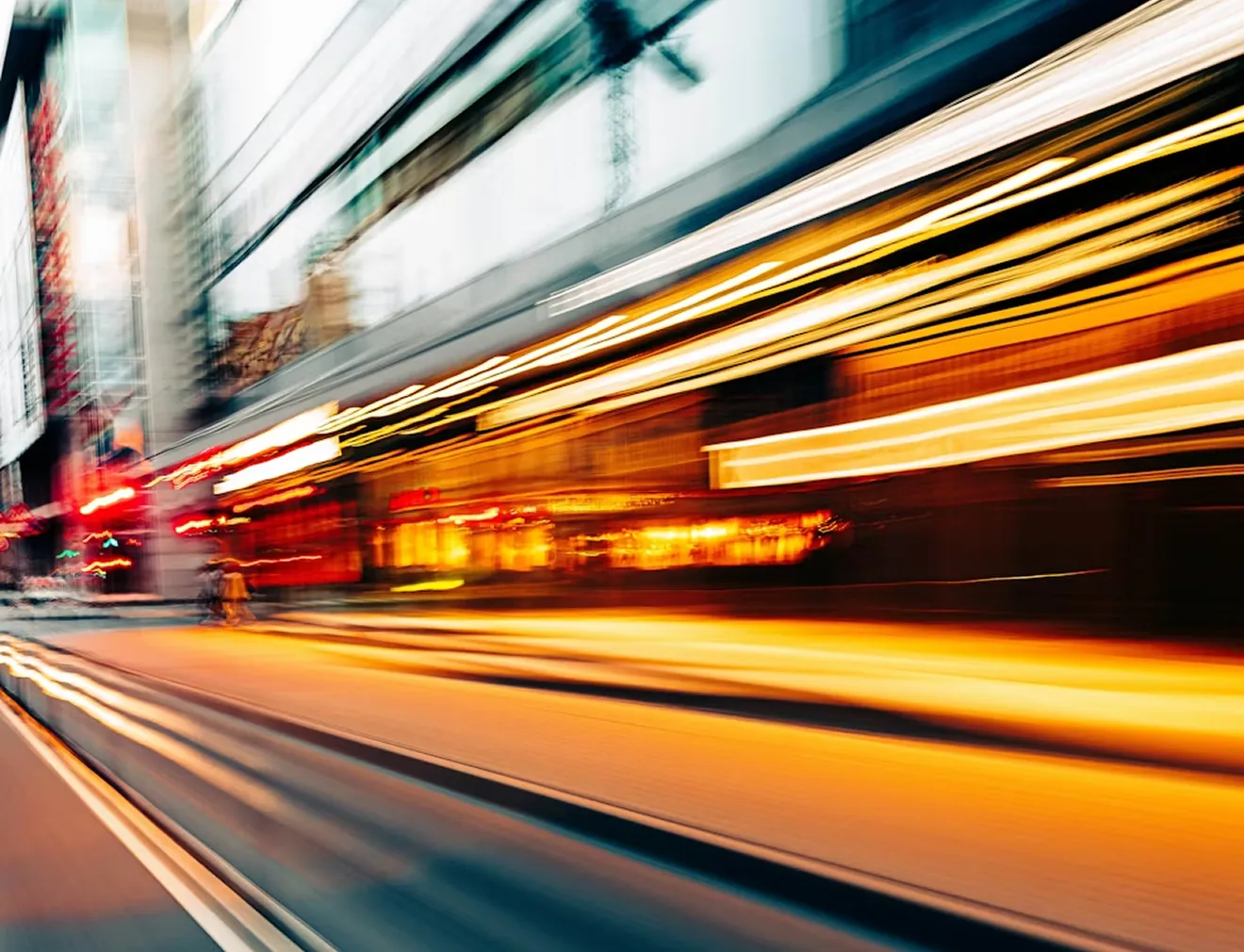 Blurred urban street scene with streaks of golden and white lights suggesting fast-moving traffic or a tram at dusk.
