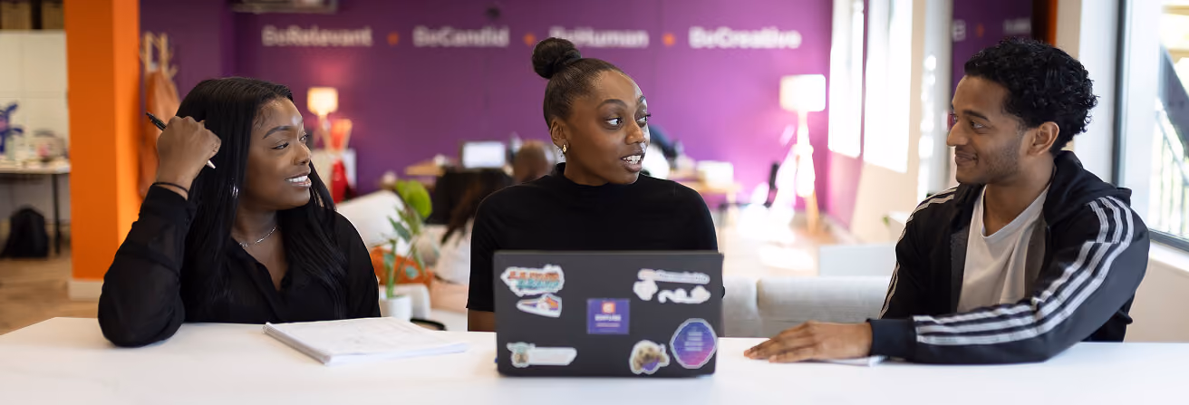 Three people sitting at a table in a bright, modern office with purple and orange walls, engaged in conversation; the person in the center has a laptop with stickers on it.