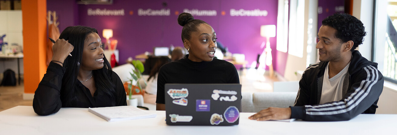 Three people sitting at a table in a bright, modern office with purple and orange walls, engaged in conversation; the person in the center has a laptop with stickers on it.
