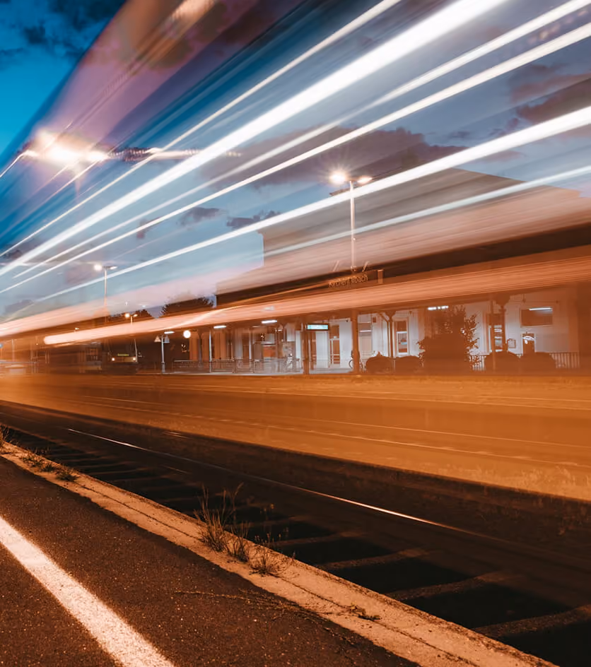 Long exposure photo of a train station at dusk with light trails from a passing train blurring across the tracks and platform.