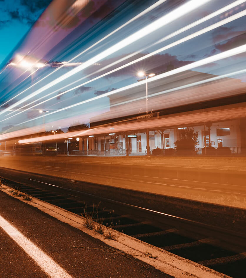 Long exposure photo of a train station at dusk with light trails from a passing train blurring across the tracks and platform.
