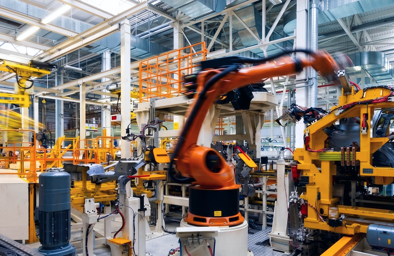 Industrial robotic arm in motion in a modern factory with various yellow and orange machinery and equipment around.
