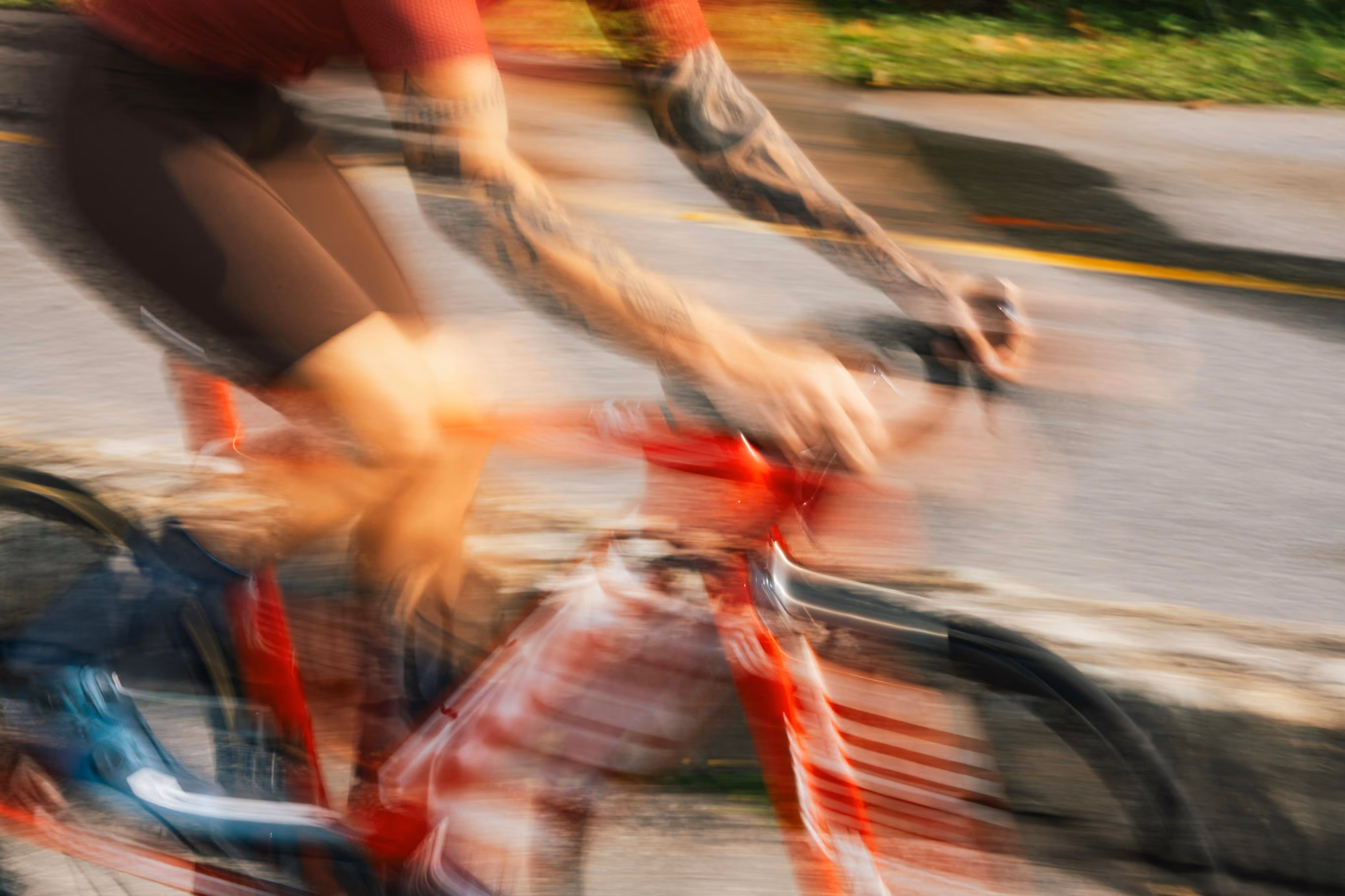 Blurred motion image of a tattooed cyclist wearing a red shirt and black shorts riding a red bicycle on a paved road with greenery in the background.
