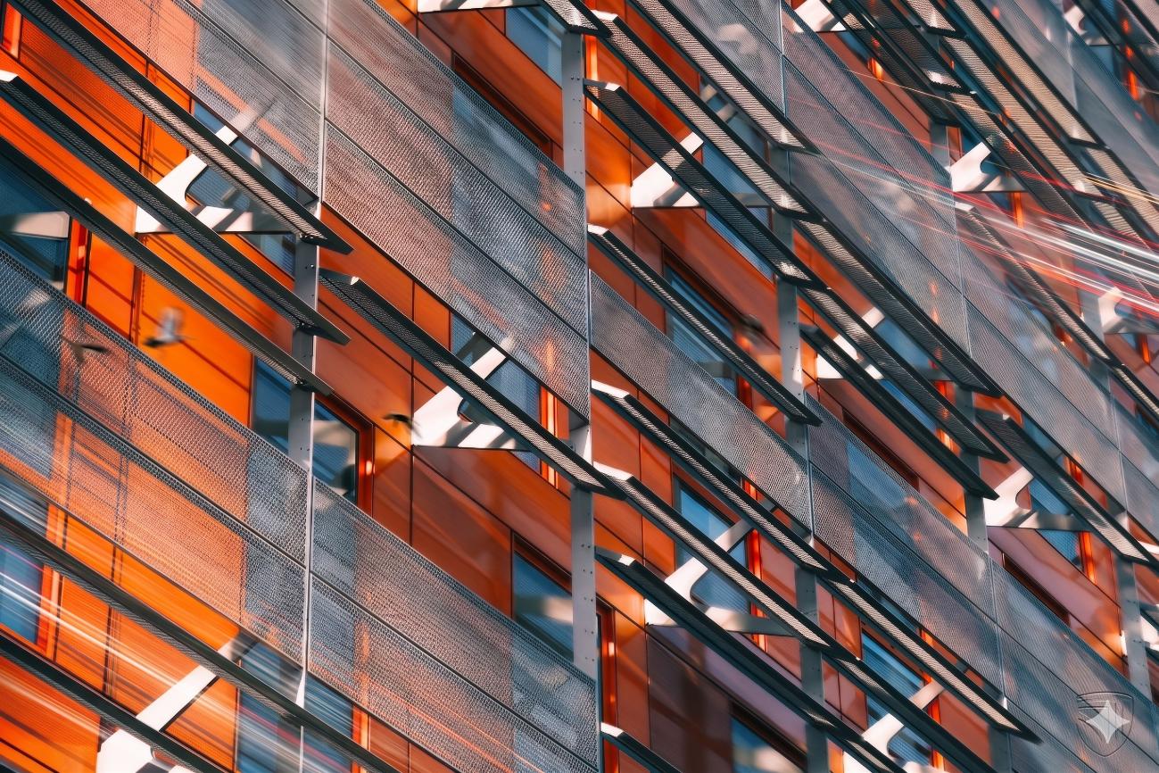 Close-up of a modern building facade featuring orange panels and metal mesh sunshades casting shadows on the structure.