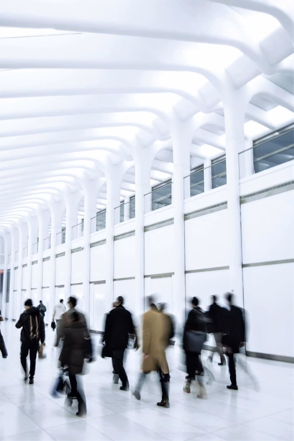 Blurred figures of people walking inside a bright, modern architectural space with tall white columns and a high ceiling with curved beams.