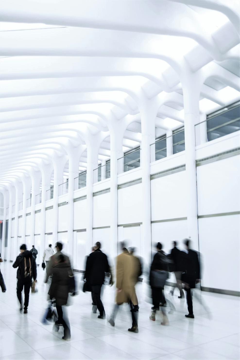 Blurred figures of people walking inside a bright, modern architectural space with tall white columns and a high ceiling with curved beams.