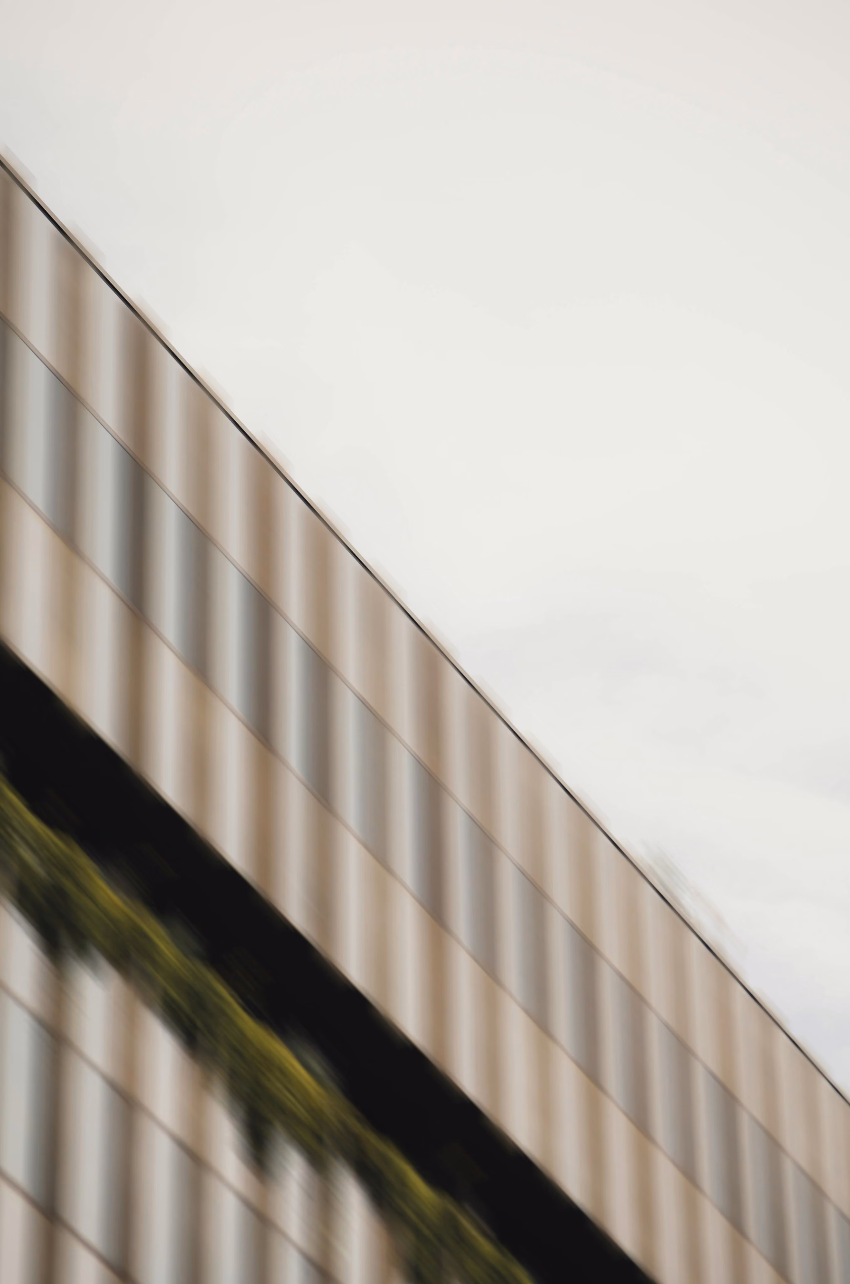 Close-up of a modern building's exterior wall with reflective glass windows and a strip of greenery, angled upwards against a pale sky.