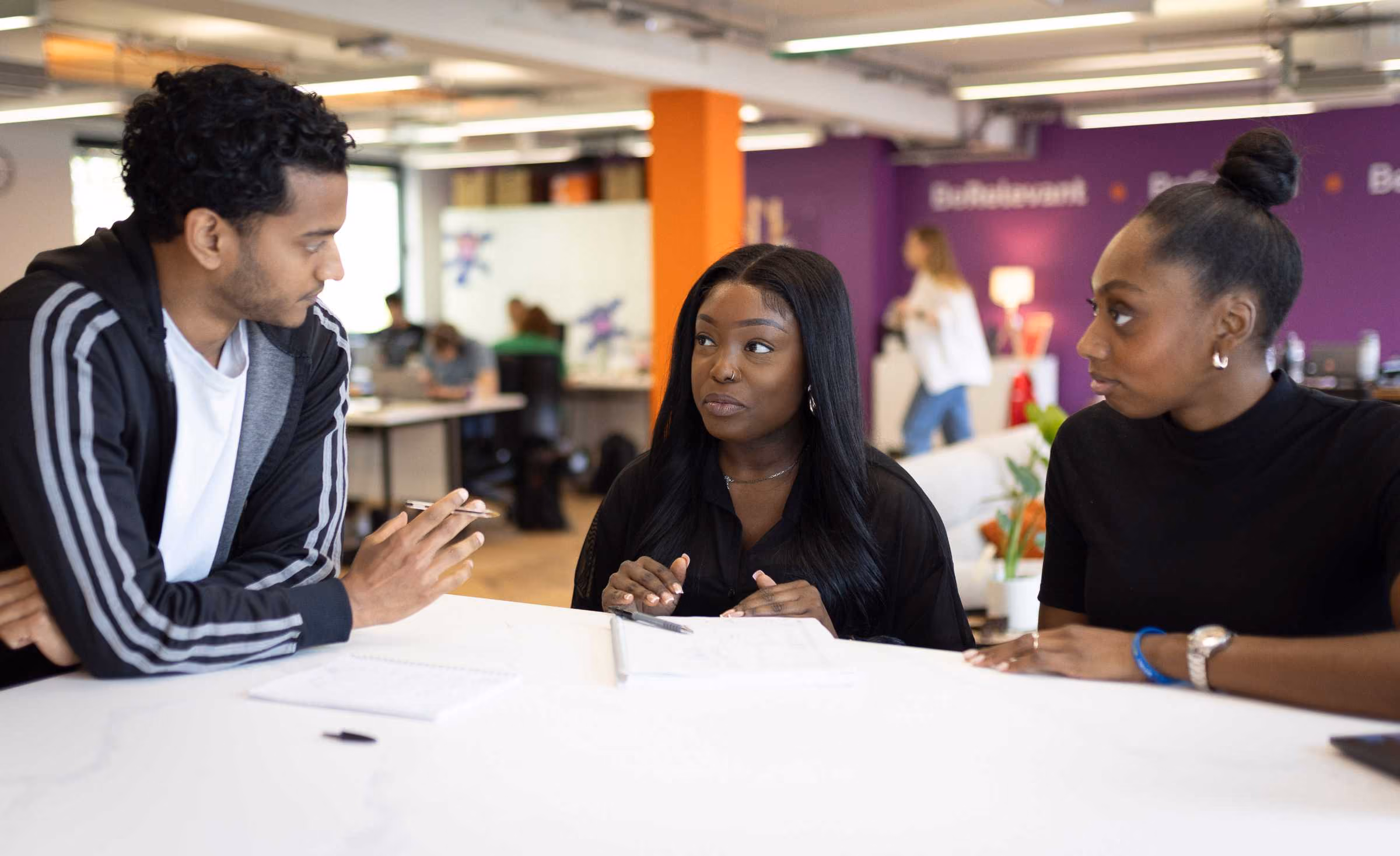 Three young adults engaged in a discussion at a table in a modern office with purple and orange walls; two women face a man who is holding a pen over notebooks on the table.