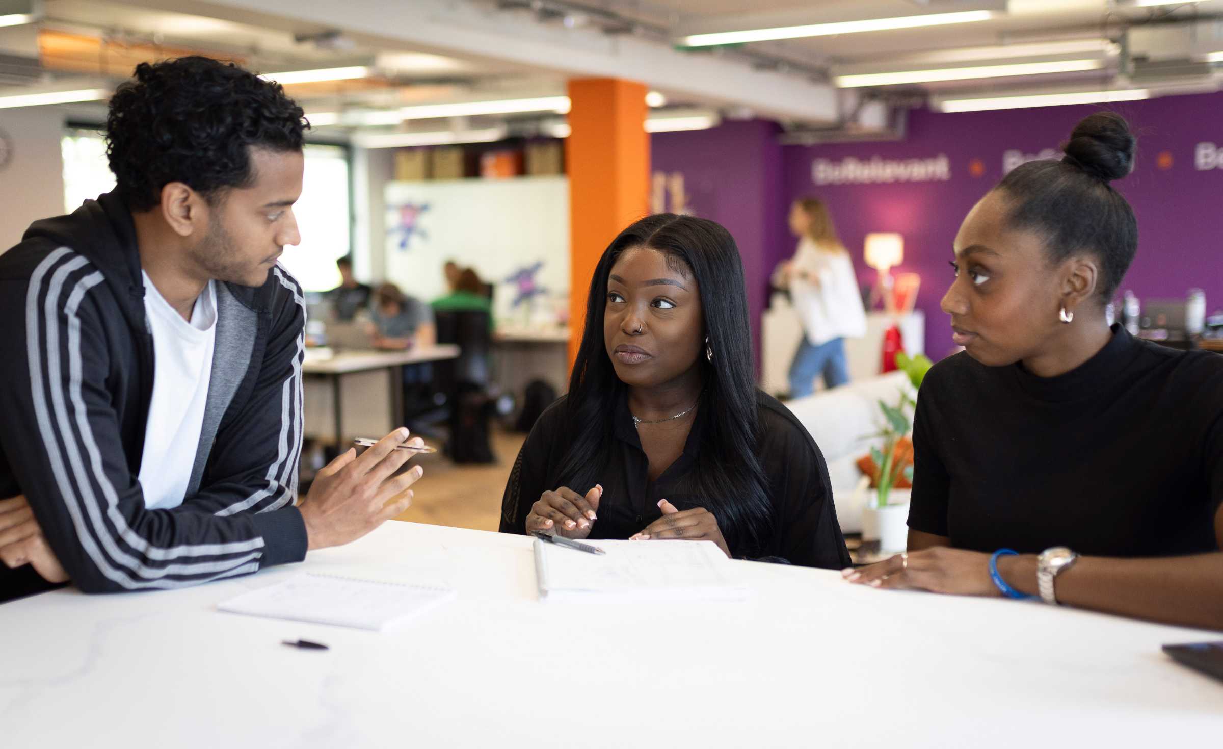 Three young adults engaged in a discussion at a table in a modern office with purple and orange walls; two women face a man who is holding a pen over notebooks on the table.