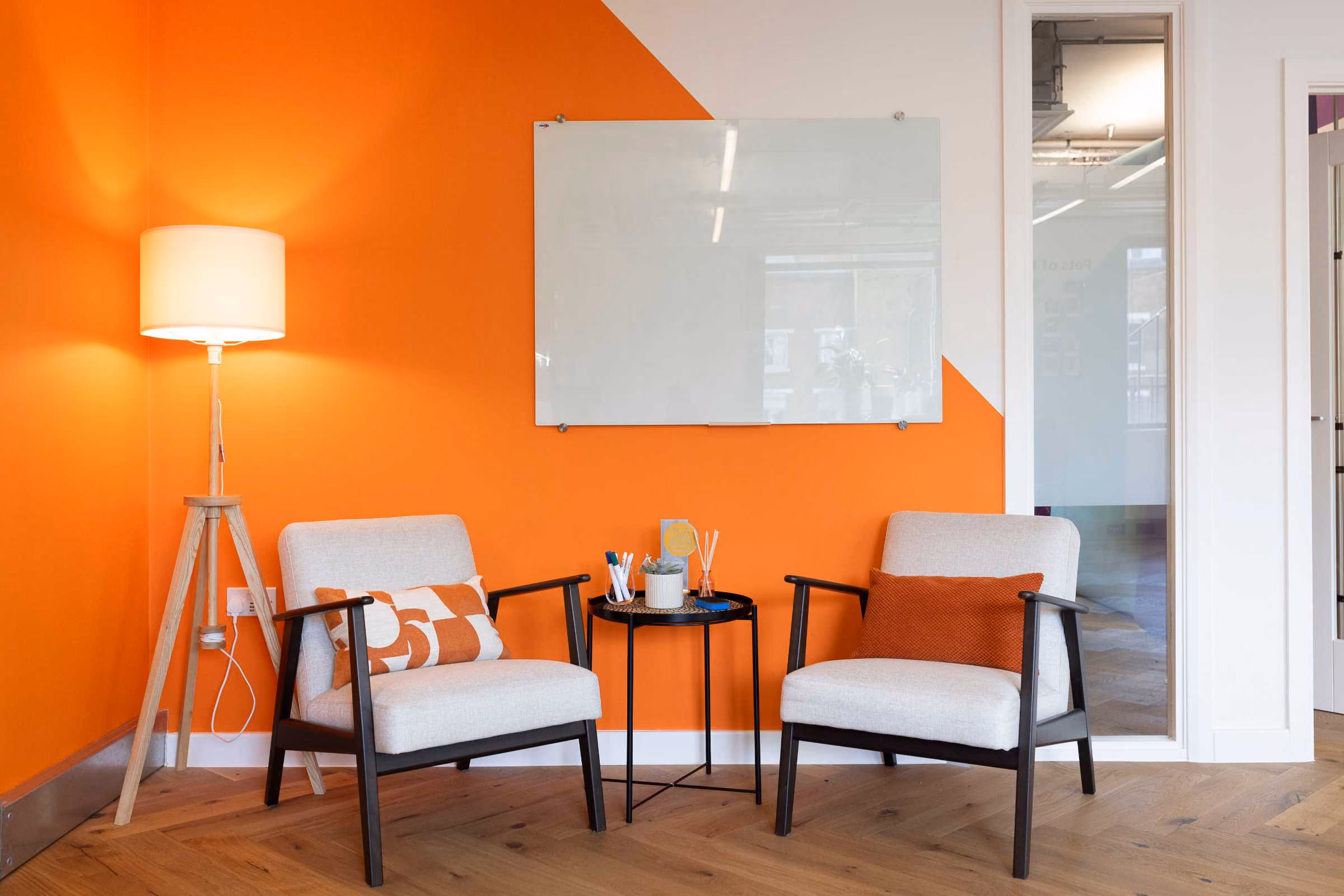 Modern office corner with two beige armchairs with orange and white pillows, a small black round table with pens and a plant, a wooden tripod floor lamp with a white shade, and an orange and white wall featuring a blank glass whiteboard.