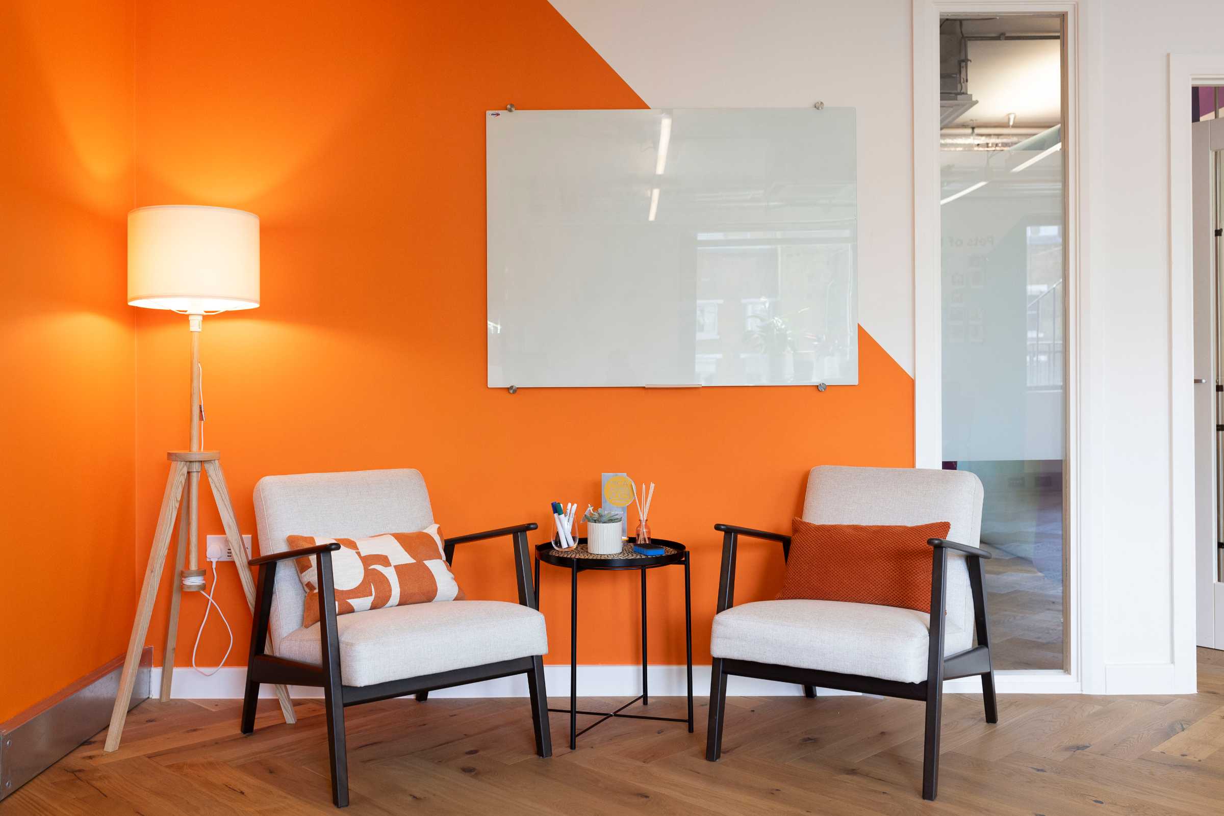 Modern office corner with two beige armchairs with orange and white pillows, a small black round table with pens and a plant, a wooden tripod floor lamp with a white shade, and an orange and white wall featuring a blank glass whiteboard.