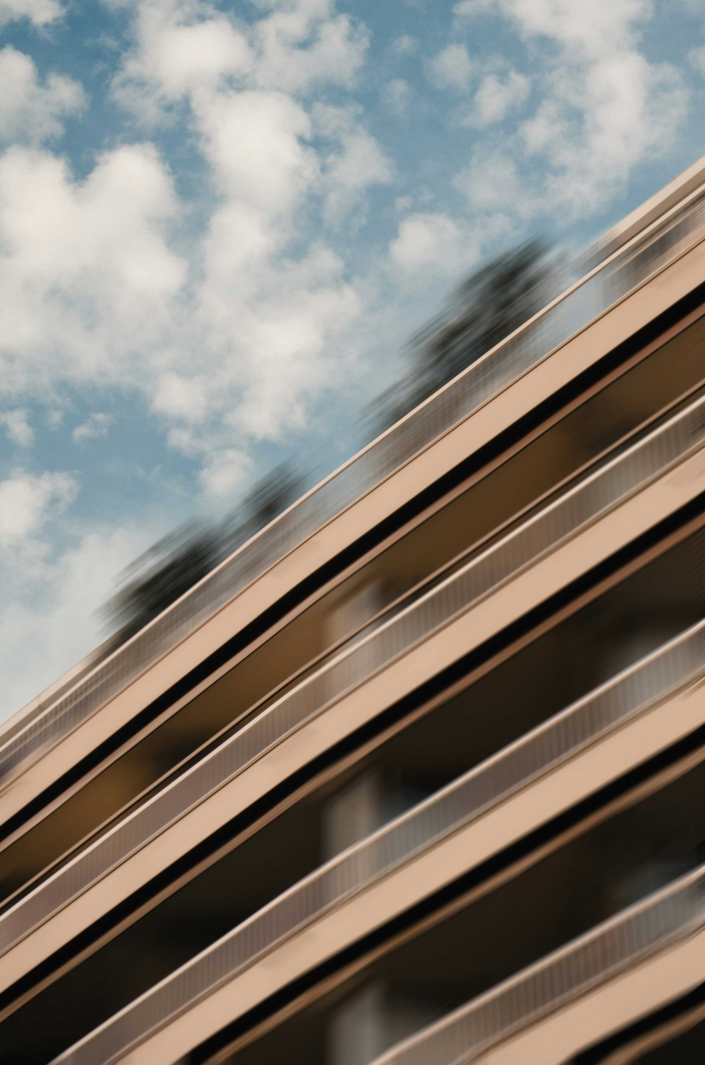Blurred close-up of a modern beige building with multiple balconies against a blue sky with scattered clouds.