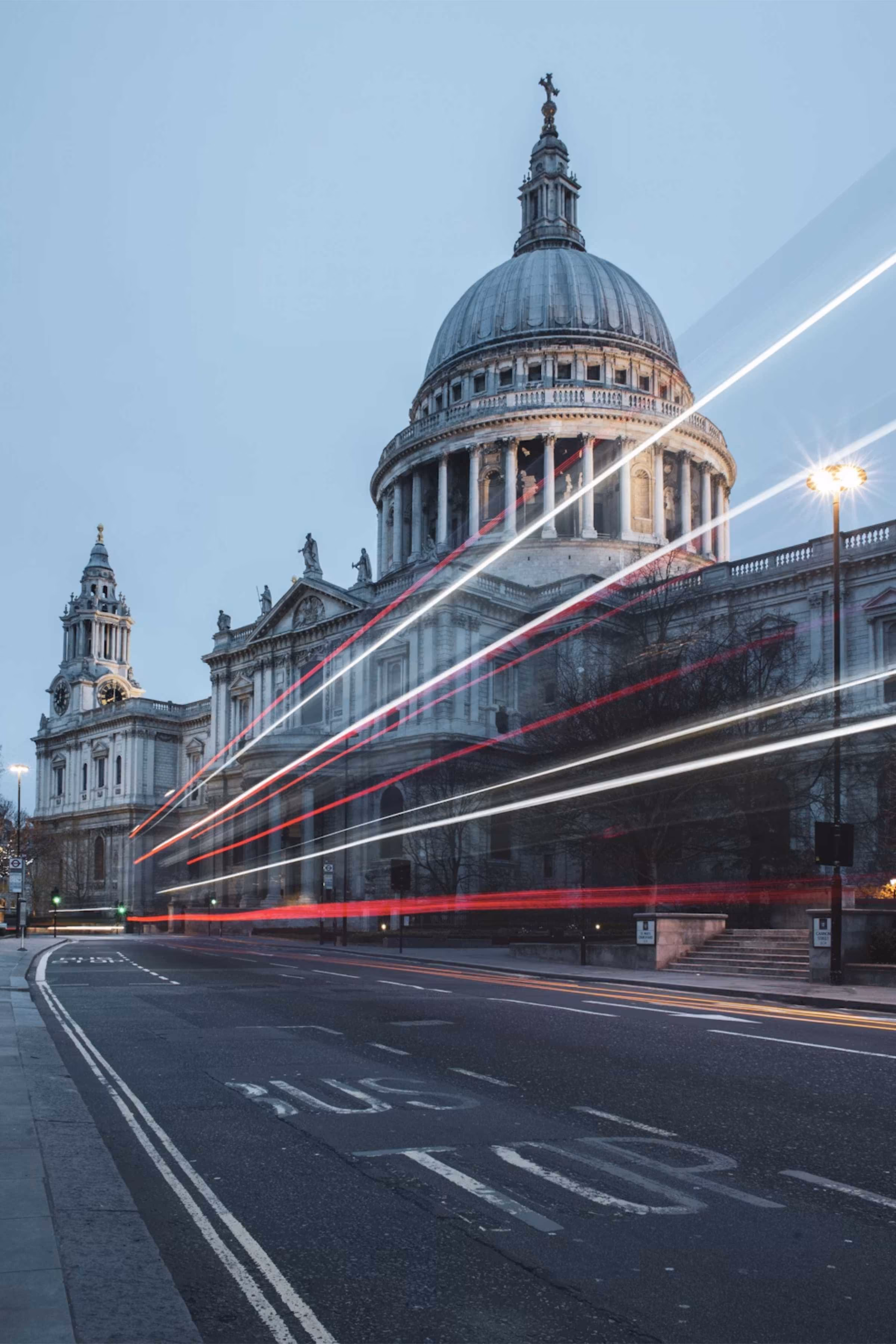 St. Paul's Cathedral in London at dusk with light trails from passing vehicles on the road in front, indicating motion and traffic.
