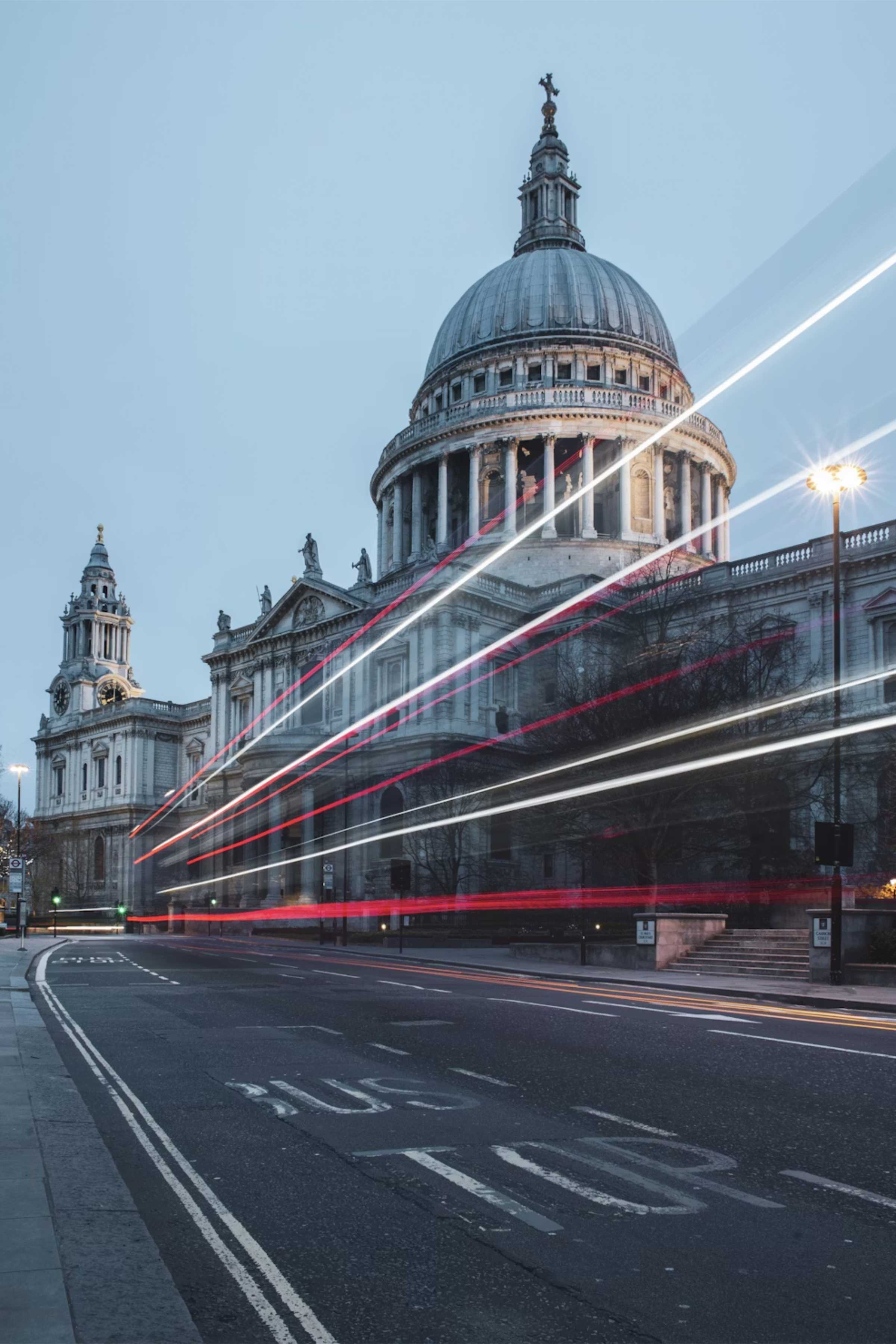 St. Paul's Cathedral in London at dusk with light trails from passing vehicles on the road in front, indicating motion and traffic.