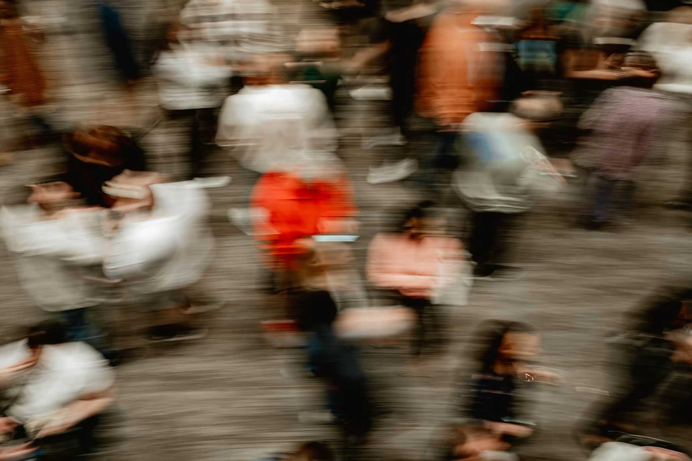Blurry motion photo of a crowded indoor space with people walking and standing, some wearing bright clothing including a notable red jacket in the center.