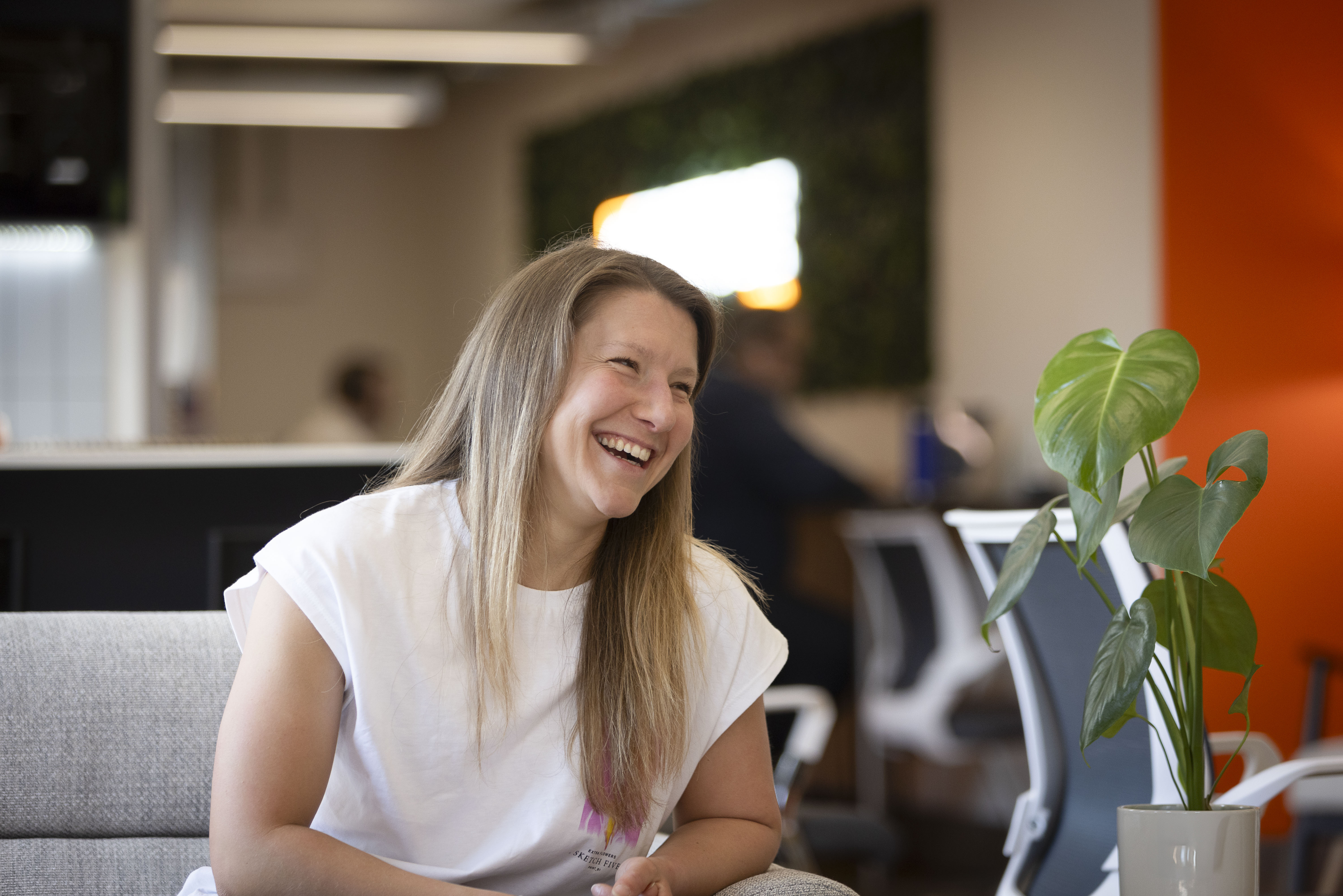 Woman with long blond hair laughing while sitting on a gray sofa in a modern office with blurred background of chairs, a bright orange wall, and a green potted plant nearby.