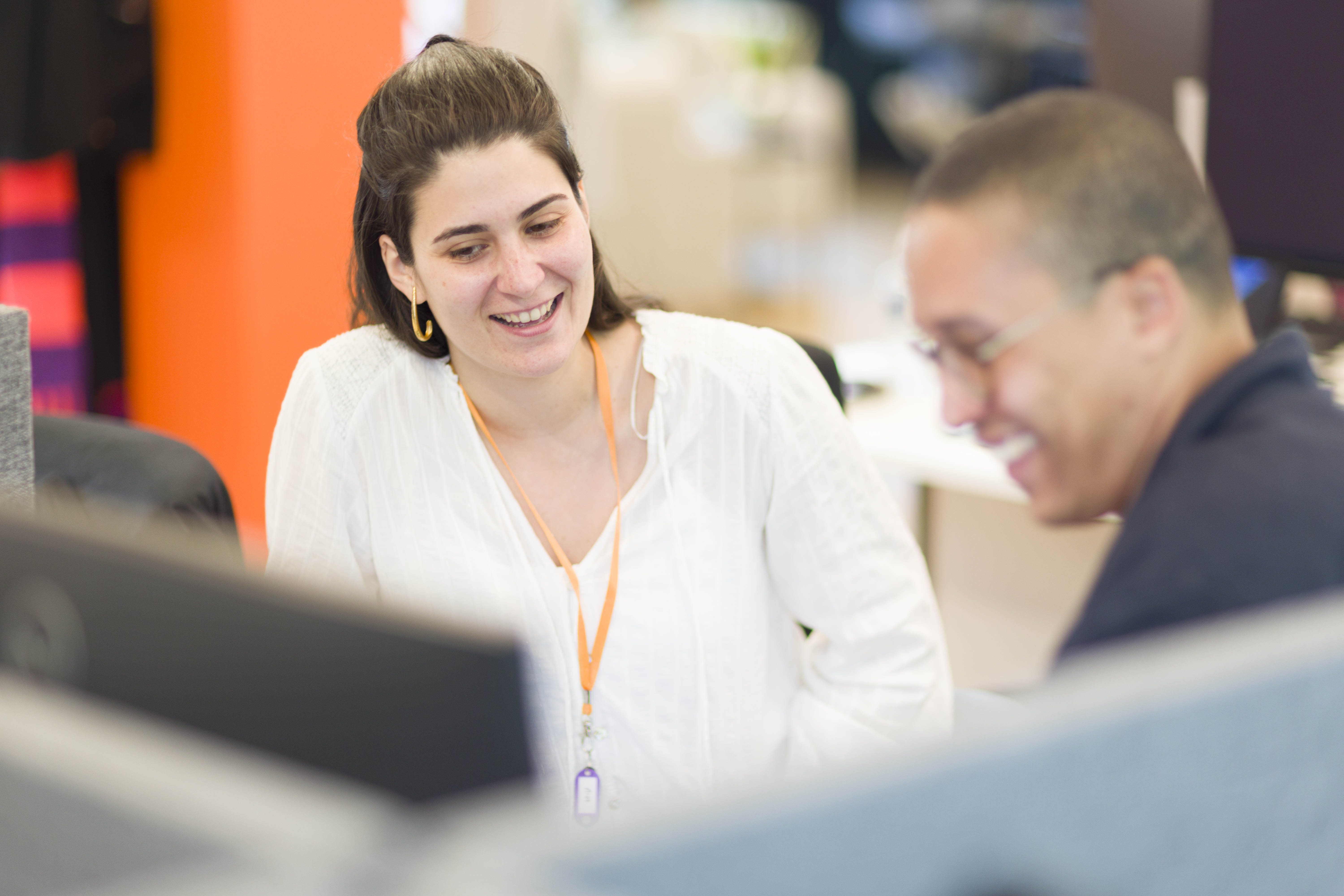 Two colleagues smiling and interacting in a bright office space, one wearing a white top with an orange lanyard and the other in glasses and a dark shirt.