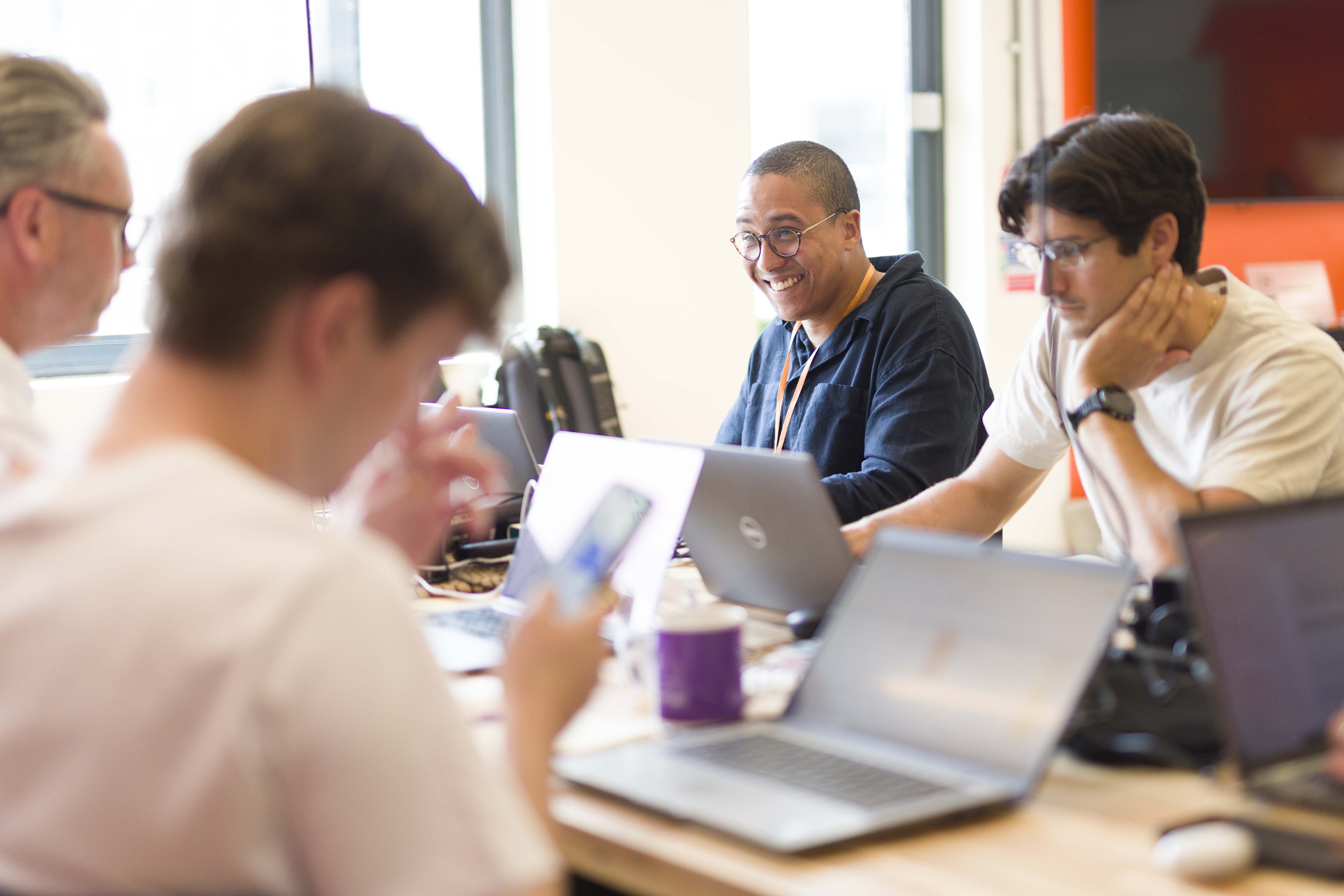 Four people working at a table with laptops and a smartphone in a bright office, one man in glasses smiling.