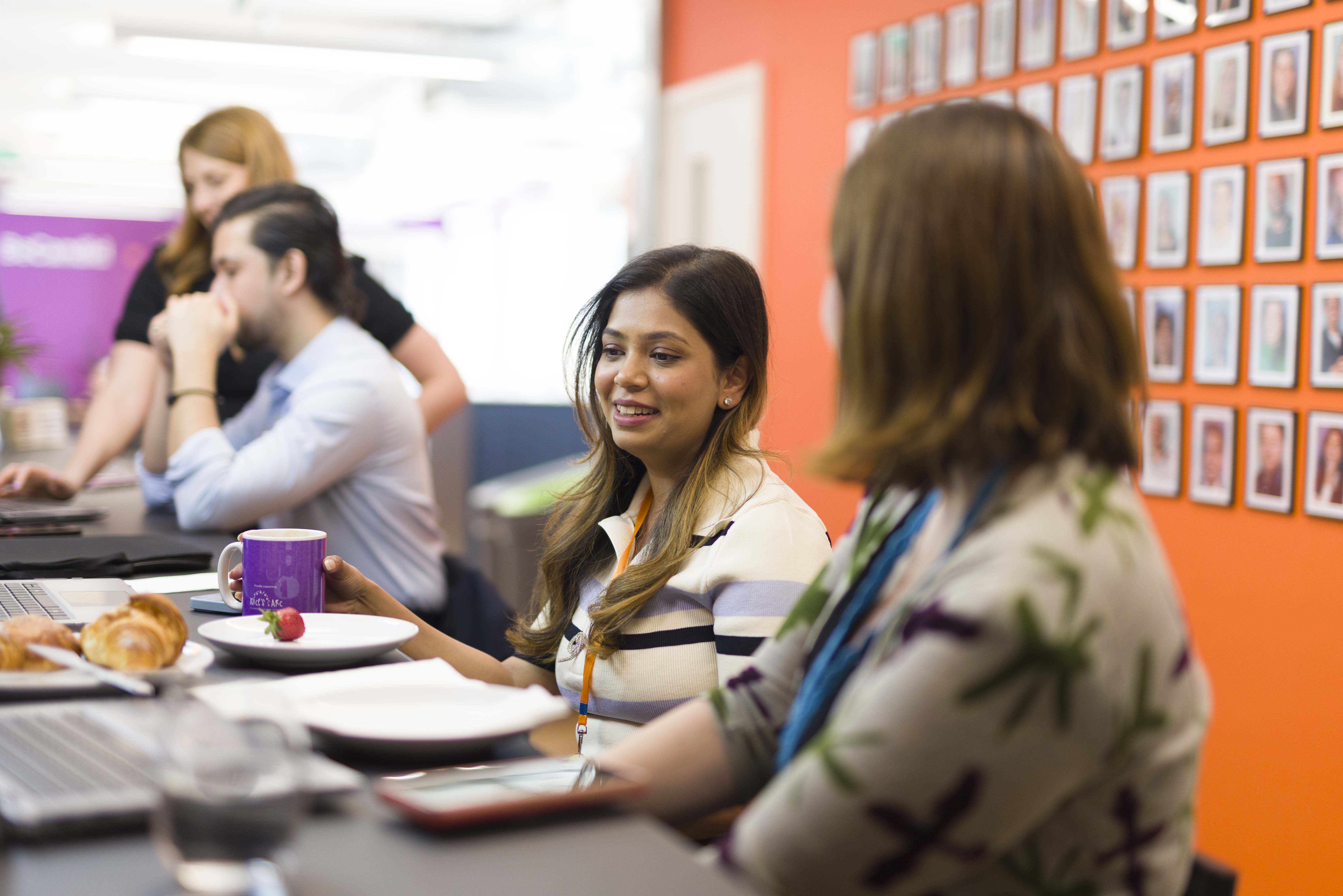 Four diverse colleagues sitting at a table in a bright office; a woman in a striped sweater holds a purple mug while talking to a woman with a floral patterned top, with a wall of framed photos behind them and a plate with croissants and a strawberry on the table.