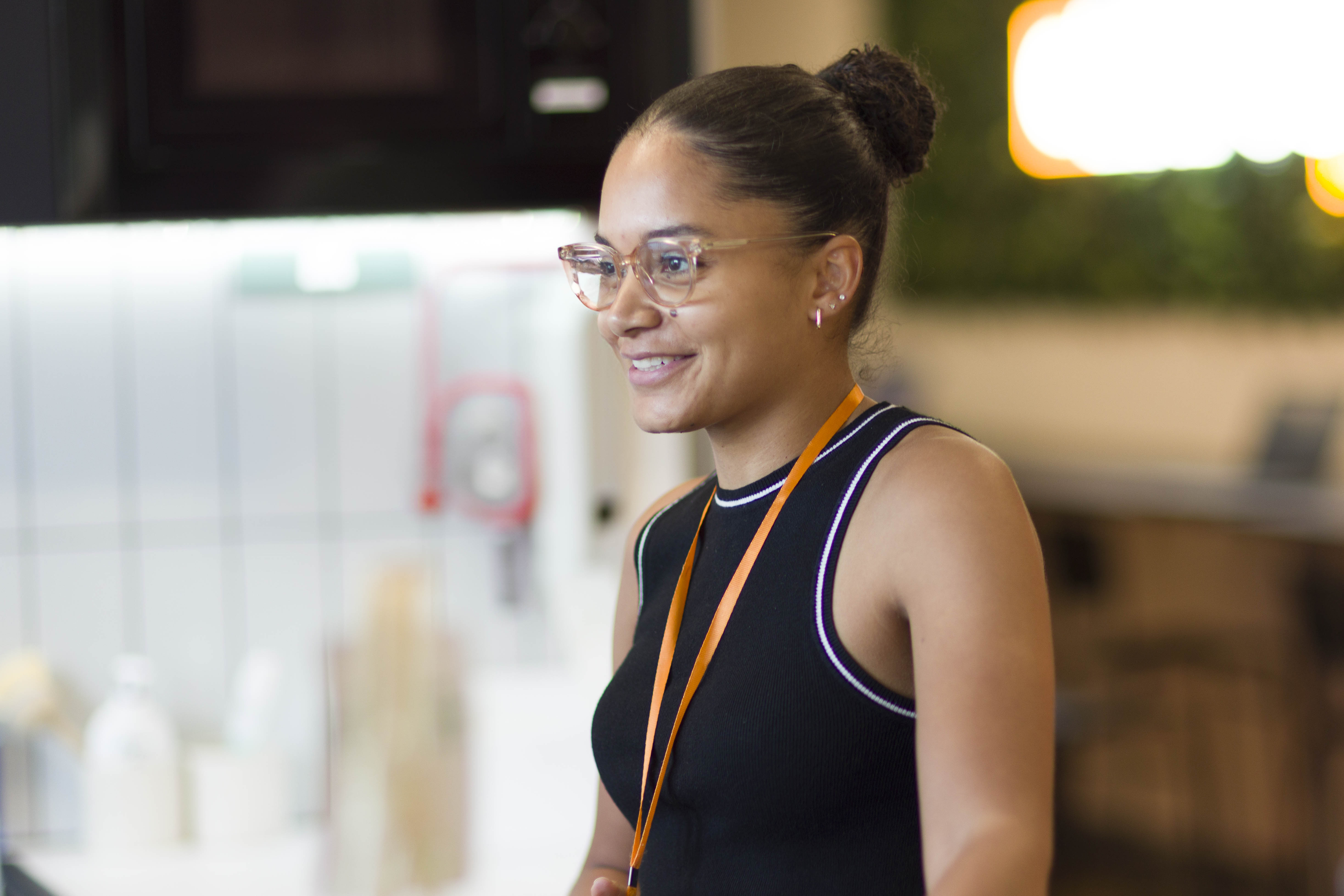Woman with glasses and hair in a bun smiling indoors, wearing a black sleeveless top and an orange lanyard.