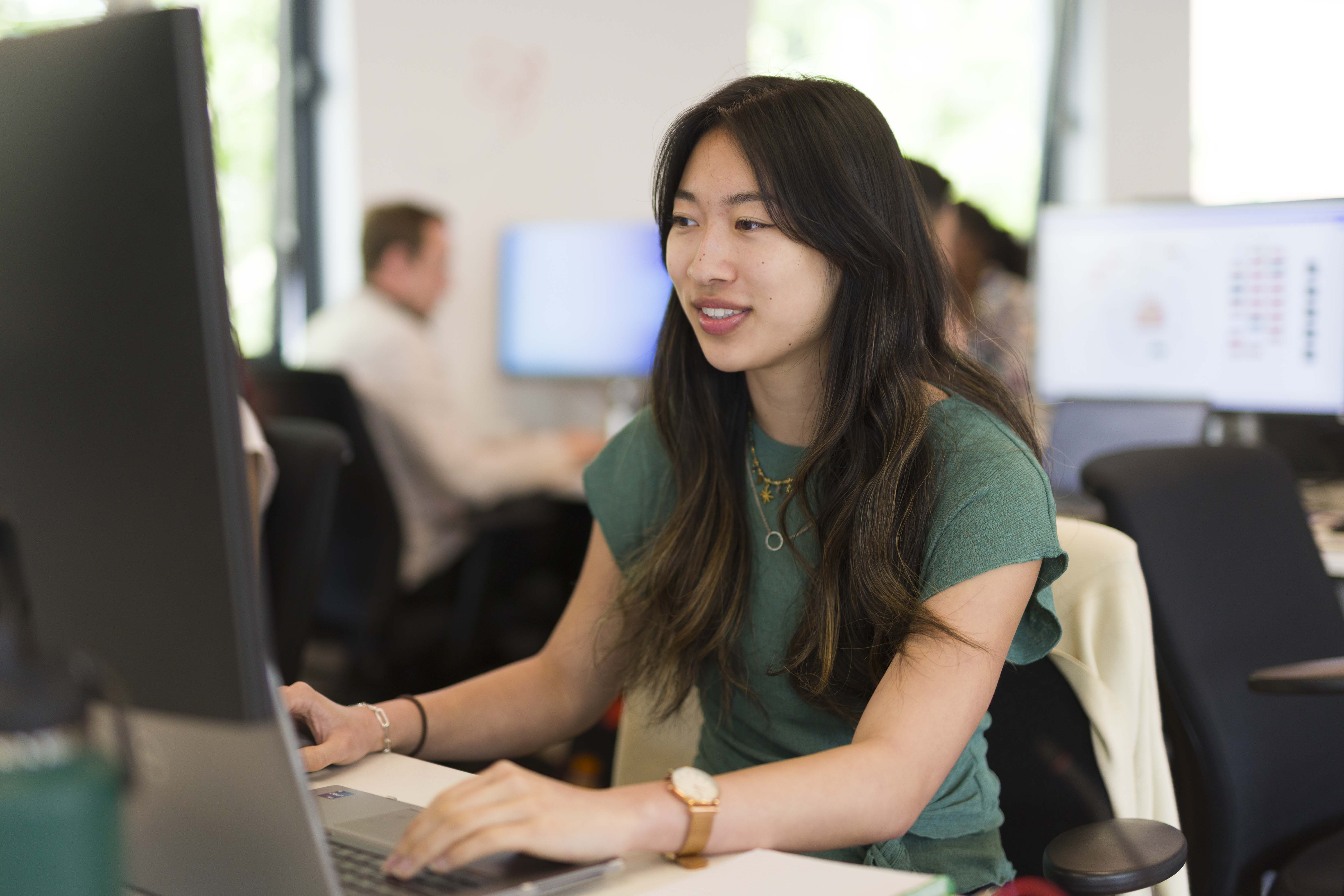 A young woman with long dark hair wearing a green shirt works on a laptop at an office desk, with blurred colleagues and computer screens in the background.