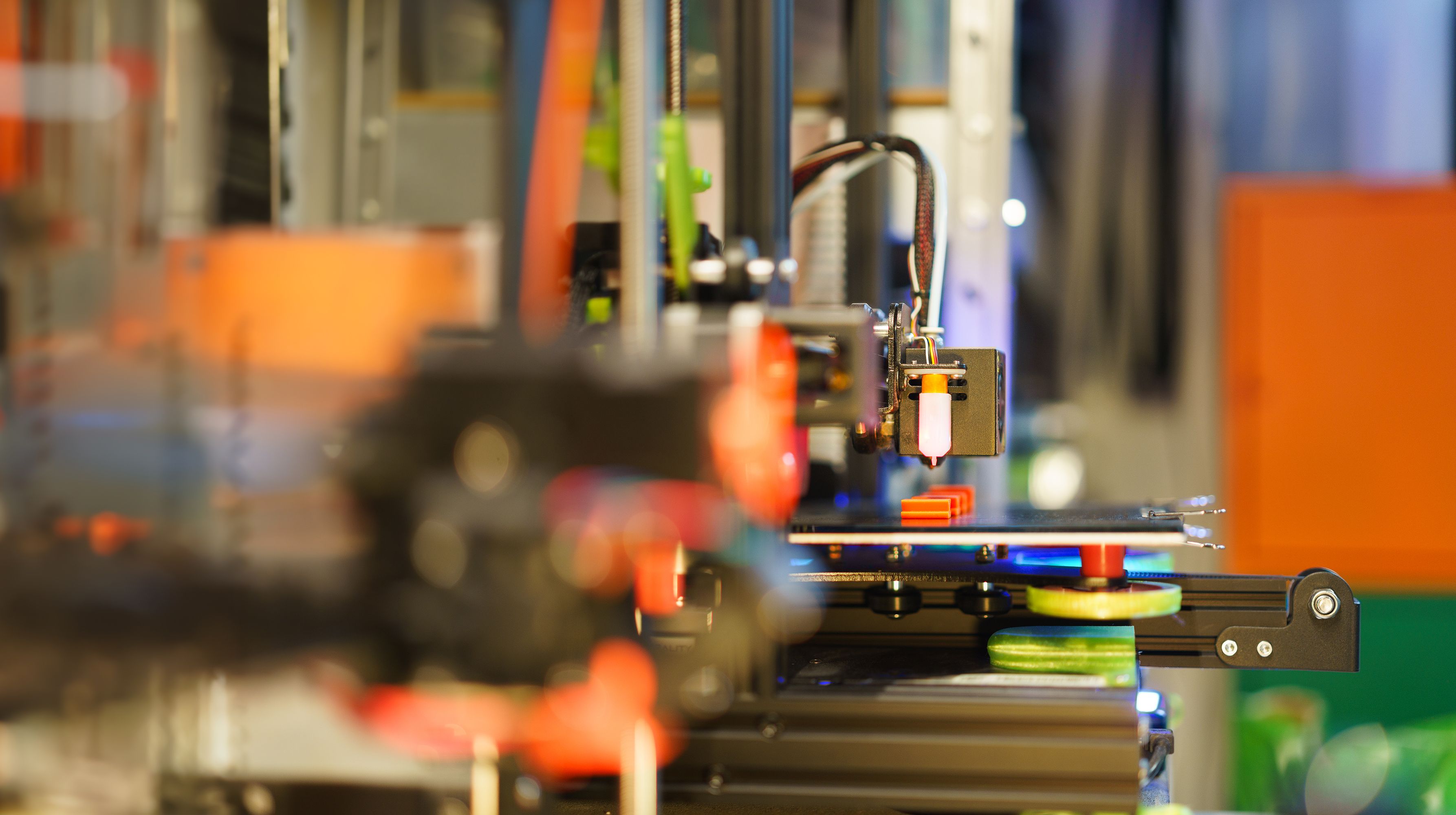 Close-up of a 3D printer nozzle actively printing orange objects on its heated bed with a blurred industrial background.