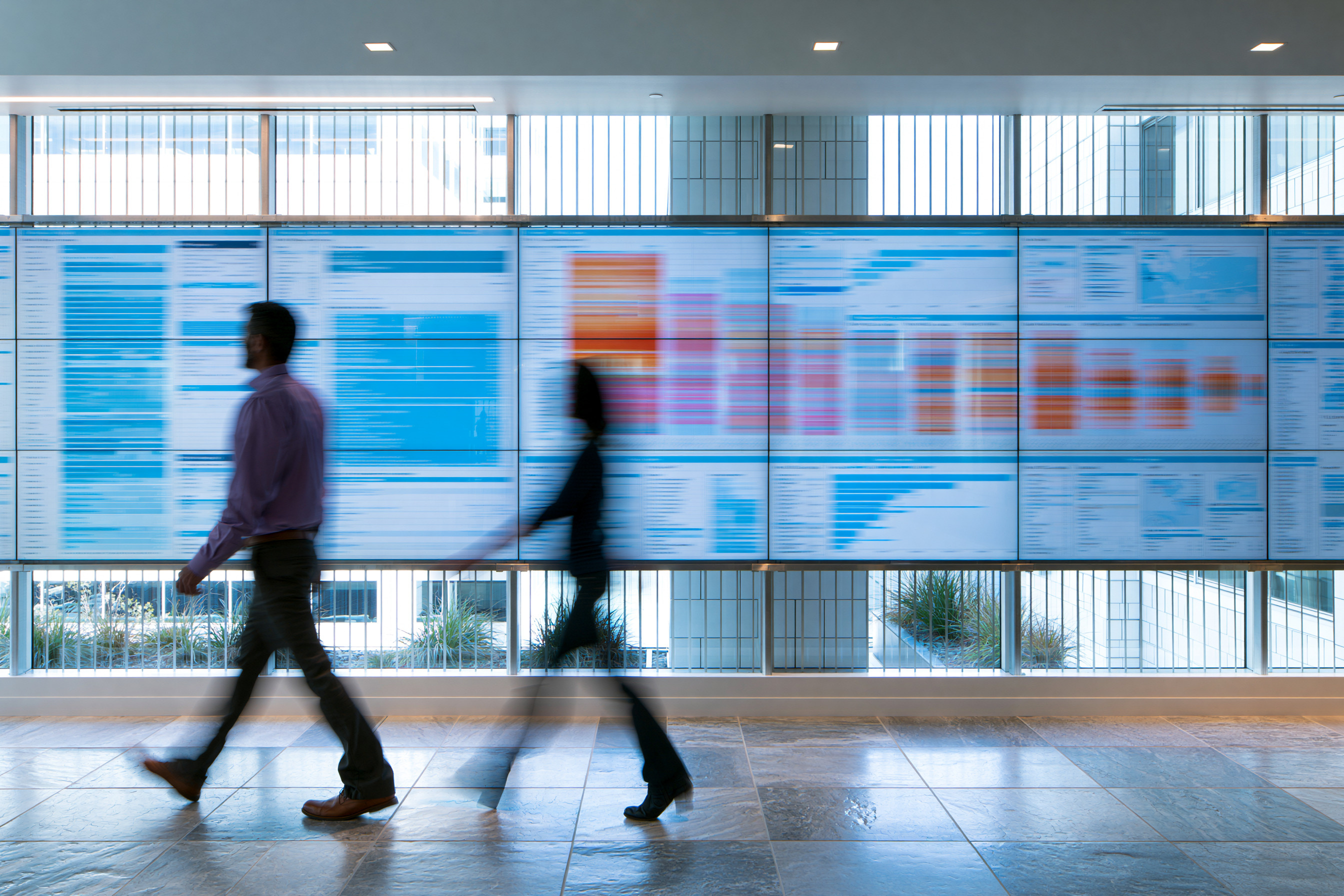 Two people walking past a large digital display wall showing blurred charts and graphs in an office corridor with large windows.