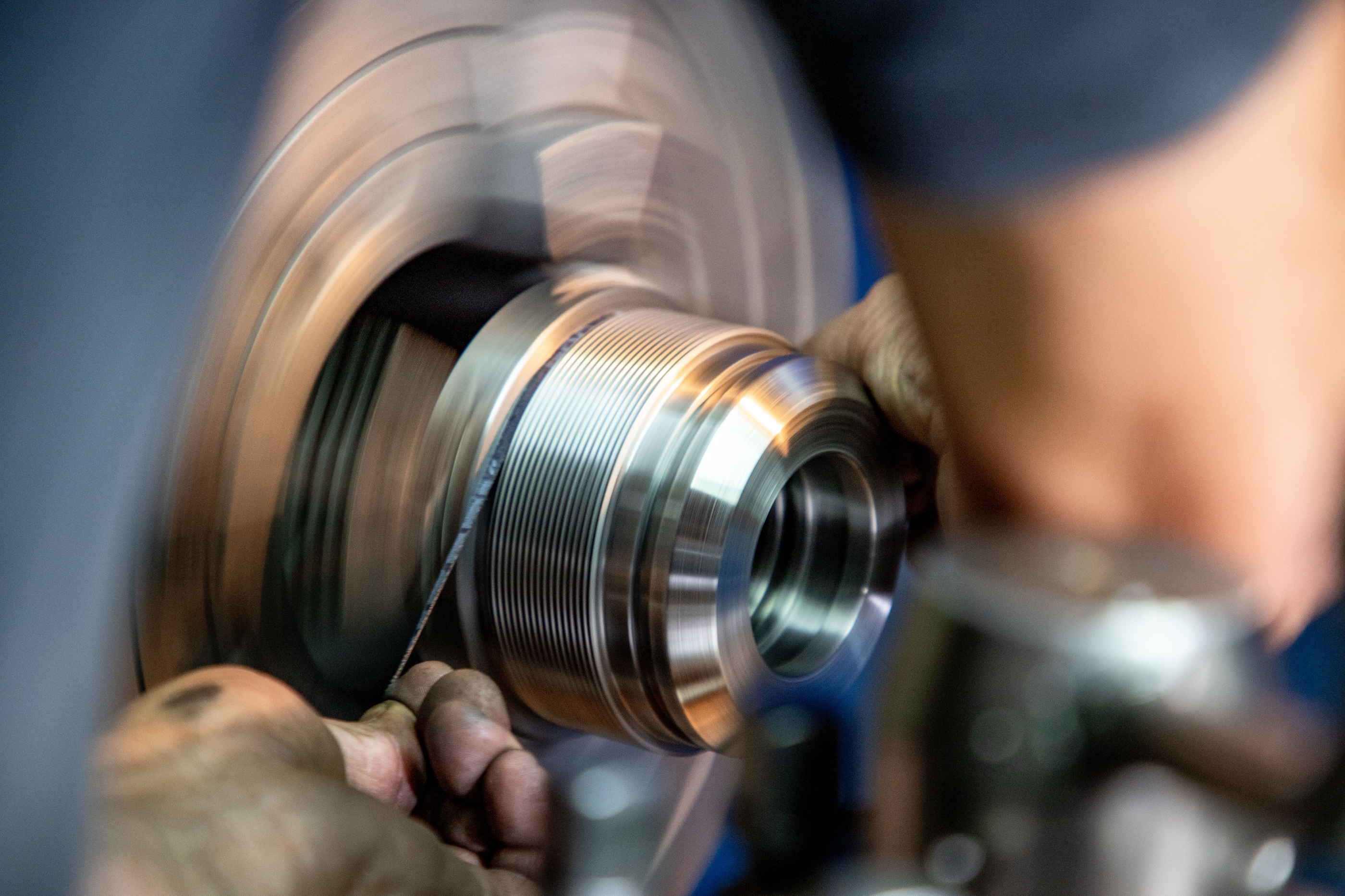 Close-up of a worker's hands holding a tool to shape a shiny, threaded metal cylindrical part on a spinning lathe machine.
