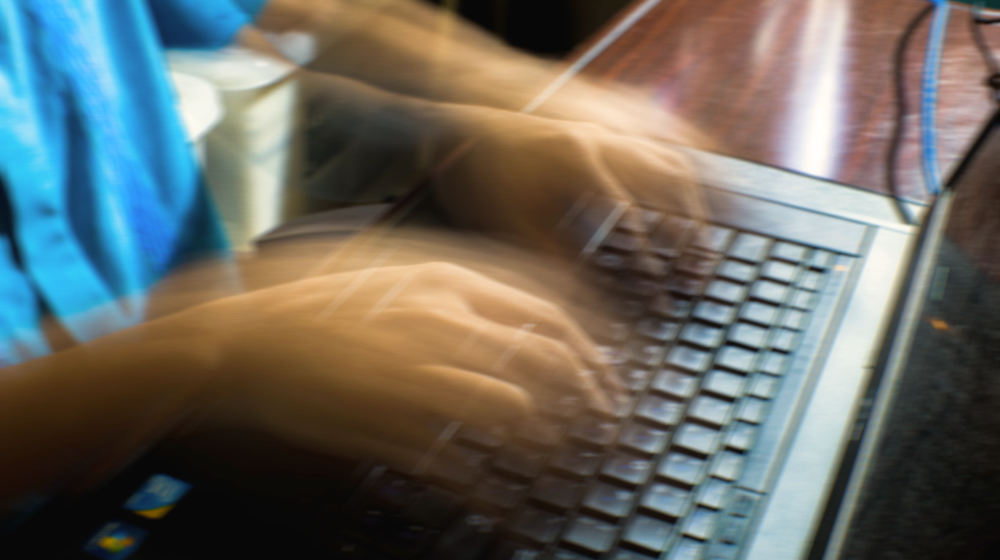Blurred motion of hands typing quickly on a laptop keyboard while wearing a blue shirt.