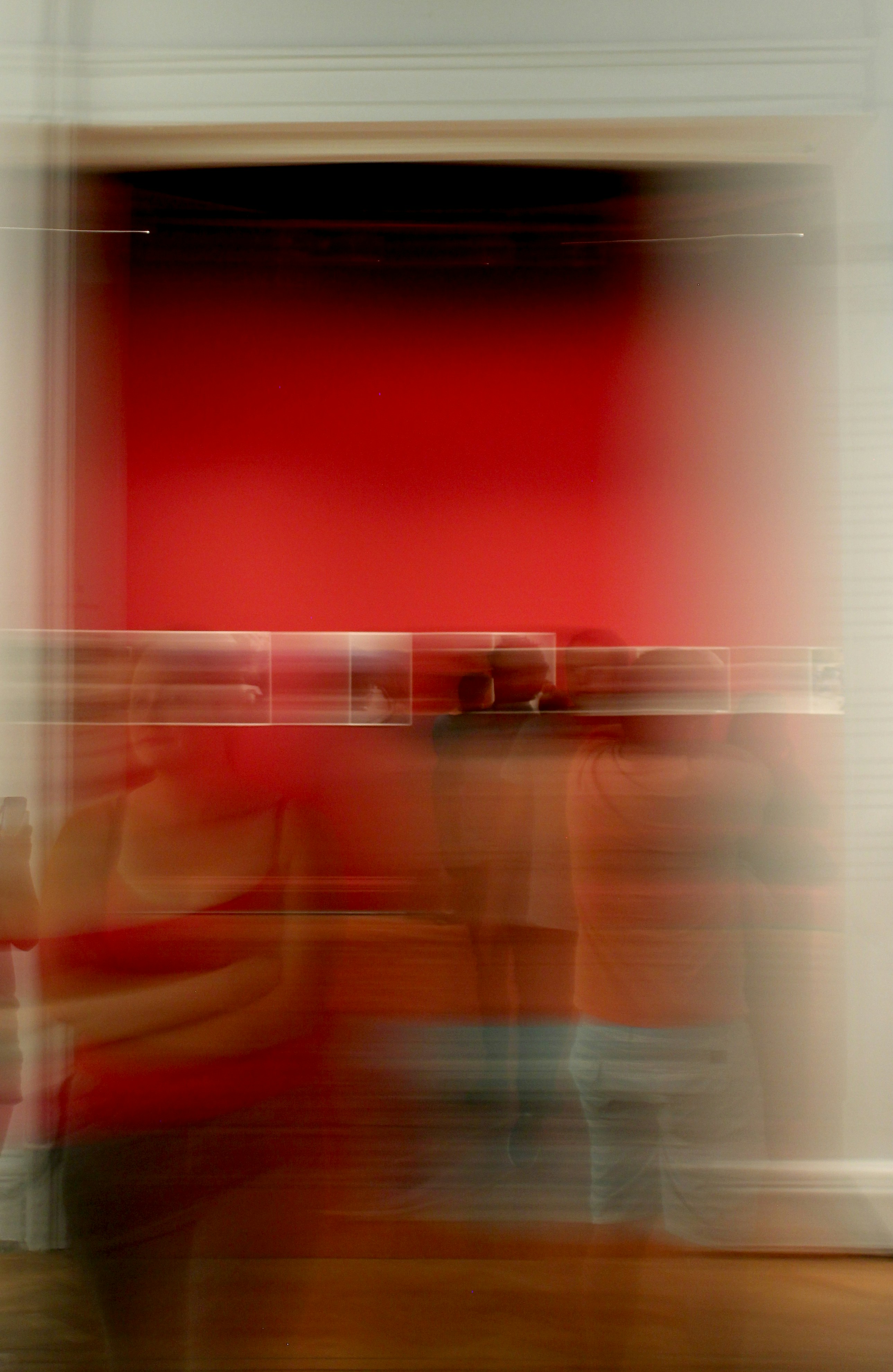Blurred image of people standing inside a building with a bright red wall featuring a horizontal line of small framed pictures or art pieces.